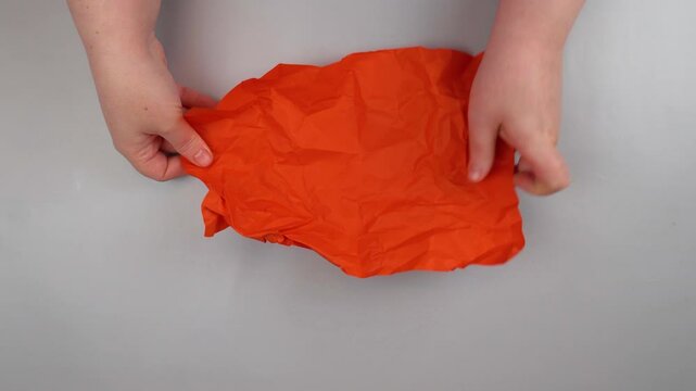 Close-up of hands spreading crumpled orange paper on a light background.   The paper has already been smoothed out a bit, but the creases are still visible.