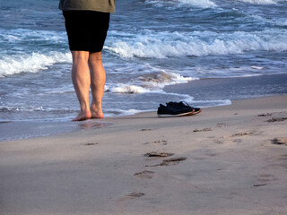 A man stands barefoot on a beach in the evening sun. His shoes lie next to him. Time spent on the...