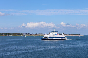 Ferry navigating through calm waters with distant shoreline and sailboats under a clear blue sky