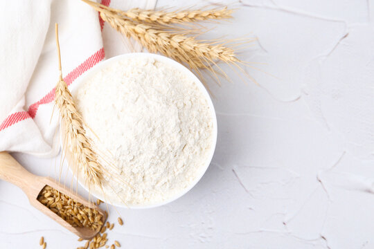Wheat flour in bowl, grains and spikes on white table, flat lay. Space for text