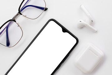 
Mobile phone with blank white screen, wireless headphones and blue light filter glasses on white...