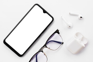 
Mobile phone with blank white screen, wireless headphones and blue light filter glasses on white background.