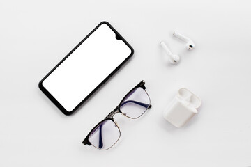 
Mobile phone with blank white screen, wireless headphones and blue light filter glasses on white...