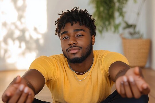 young man with curly hair and beard sitting on the floor wearing a yellow t-shirt reaching forward with both hands in a bright room with blurred plants in the background