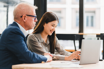 Young woman and senior man working together on laptop in modern office, smiling and focused on screen, collaborative business meeting with diverse colleagues