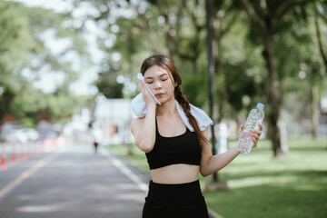 Young Asian woman drinking water after exercise, standing outdoors in workout clothes with towel,...
