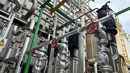  Industrial cooling tower system emitting vapor under blue sky. Heavy-duty piping, valves, and heat exchangers in a manufacturing or refinery plant exterior.