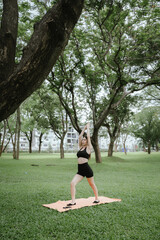 A young woman practices yoga outdoors in a peaceful urban park, promoting wellness, balance, and mindfulness through stretching exercises on a mat surrounded by nature and tranquility.
