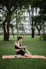 A young woman practices yoga outdoors in a peaceful urban park, promoting wellness, balance, and mindfulness through stretching exercises on a mat surrounded by nature and tranquility.
