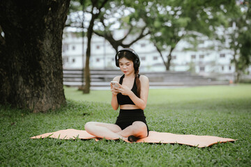 Young Asian woman sits in a quiet park. She wears headphones, enjoys music, practices mindfulness, exercises and digital health with her smartphone.