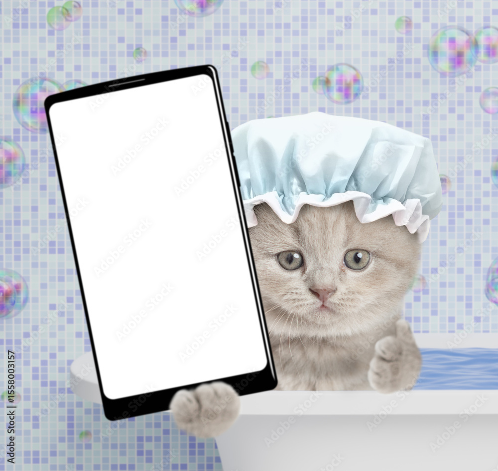 Poster cute kitten wearing shower cap takes the bath at home and shows big smartphone with white blank scre