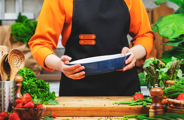 Woman chef holds a ceramic baking dish for cooking healthy meals in a warm kitchen surrounded by fresh vegetables and herbs