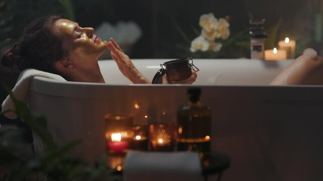 A serene woman in her 30s, adorned with a gold face mask, gently touches her cheek while relaxing in a candlelit bathtub, surrounded by lush plants, creating a luxurious beauty and wellness experience