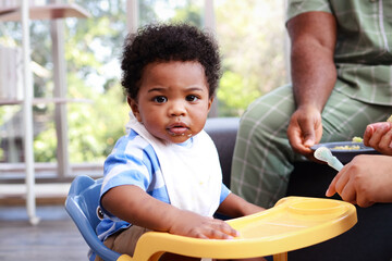 Portrait of a black boy sitting at the dining table waiting for his mother to feed him in the living room of his home. Preschool child development. Menu for 1 year old baby.