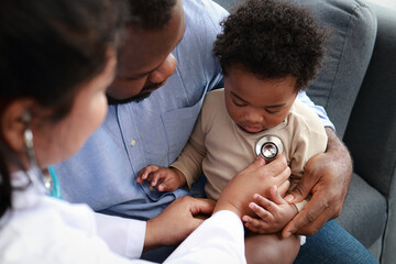 Doctor uses stethoscope to examine heart of little boy sitting on father's lap to check for possible abnormalities in child. Child health concept. Medical services.