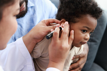 Doctor uses flashlight to examine the ears of little boy sitting on his father's lap to check his...