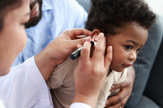 Doctor uses flashlight to examine the ears of little boy sitting on his father's lap to check his hearing and detect possible abnormalities in children. Child health concept.