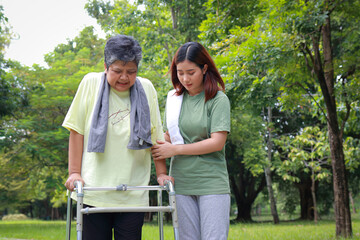 An elderly Asian woman walks with a cane in an outdoor park, with her daughter or a physical therapist taking care of her. Health care center for the elderly.