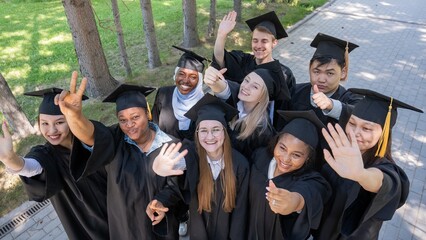 Students of different nationalities in graduation gowns wave their hands. 