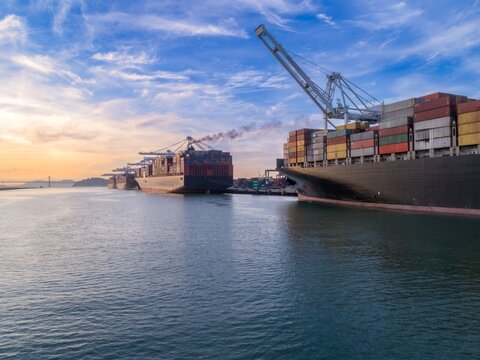 Cargo ships are docked at the Port of Oakland in California, USA. Cranes are loading and unloading shipping containers. The ships transport goods globally.