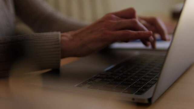 An elderly woman's hands are shown typing on a laptop keyboard at home, suggesting themes of aging, technology, and remote work.