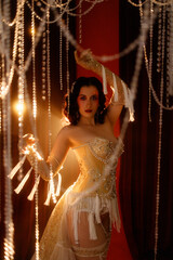 Brunette young woman, Caucasian, dressed as vintage cabaret dancer, posing front view under hanging beads on dark background. Concepts: cabaret, retro glamour, burlesque, stage show.