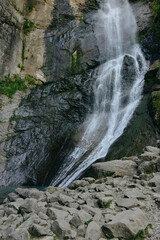 Natural waterfall cascading down rocky cliff surrounded by green moss and rugged stones under...