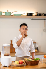 Asian man closing eyes enjoy drinking a glass of protein smoothie after making it at kitchen counter