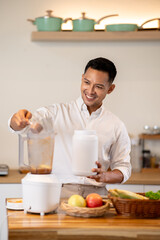 Asian man adding whey protein powder into blender while making smoothie at kitchen wooden counter.