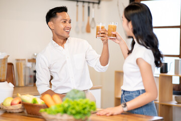 Asian man and woman clinking glass of smoothie after making drink together at kitchen wooden counter