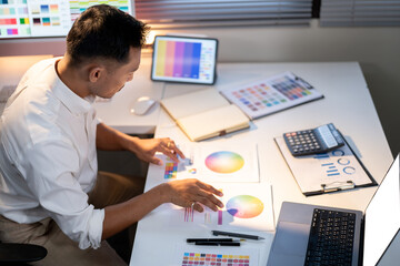 Asian man designer looking at color palettes while sitting with laptop and tablet on working table.