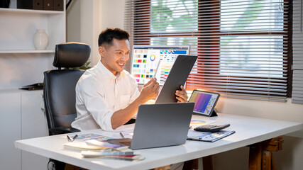 Happy asian man designer holding clipboard looking at stylus while sits at office's working table.