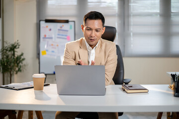 Asian businessman excitedly talking or explaining on video chat via laptop at office's working...