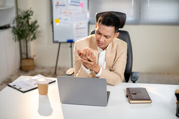 Asian businessman holding his strained wrist after working too long on laptop at office's table.