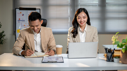 Asian businessman writing in notebook as female coworker looking at laptop sitting at office's table.