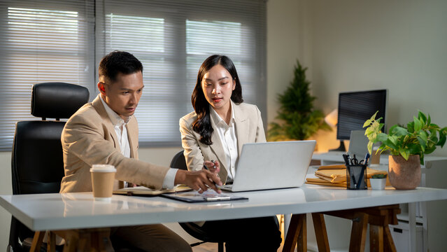 Asian businessman pointing at clipboard to female coworker discussing aside laptop at office's table