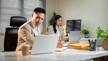 Businessman writing with pen in notebook and looking at laptop on office table aside female coworker
