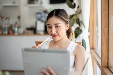 Asian woman holding stylus touching lips looking at tablet sitting at table aside cafe's glass wall.