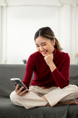 Pretty asian woman smiling as looking at phone fist under chin while sitting on sofa in living room.