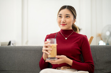 Pretty asian woman smiling and holding iced coffee while sitting on sofa in a living room or lounge.