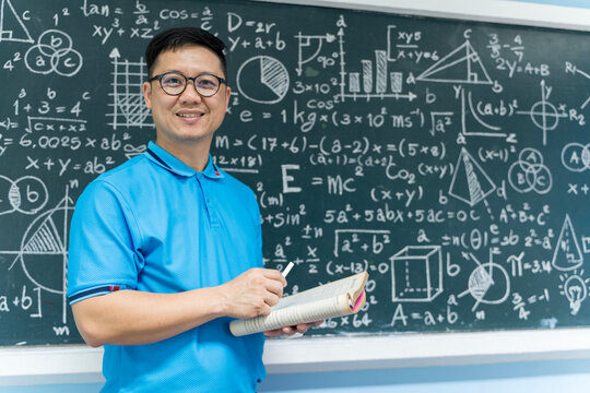 Asian male teacher holding book and chalk standing in front of math blackboard, education and academic concept
