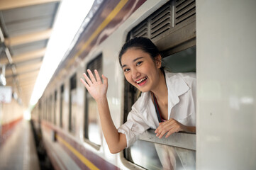 Smiling asian woman leaning head and waving hand out of carriage's window at train station platform
