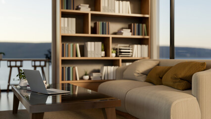 Laptop and books on a glass top wooden table beside sofa and bookshelf in a living room or library.