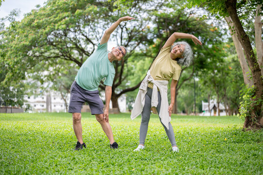 Glasses old man and old woman smiling while standing and warming up together on grass lawn in park.