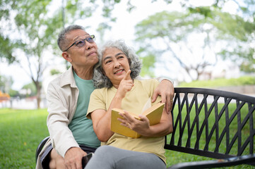 Fototapeta premium Old woman holding book and pointing as hugged by glasses man while sitting together on bench in park