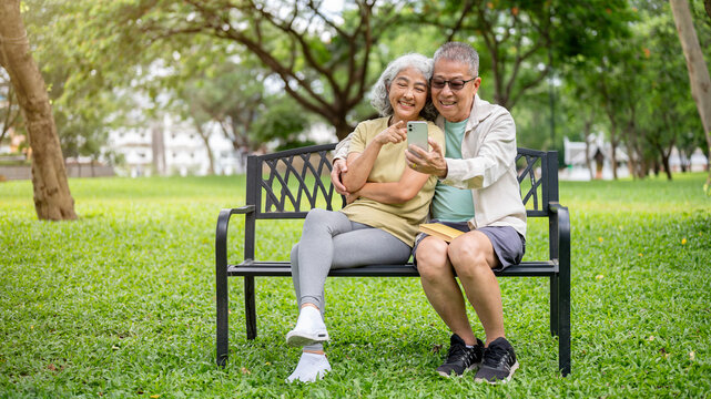 Glasses old man holding phone taking photo watching video with woman while sitting on bench in park.