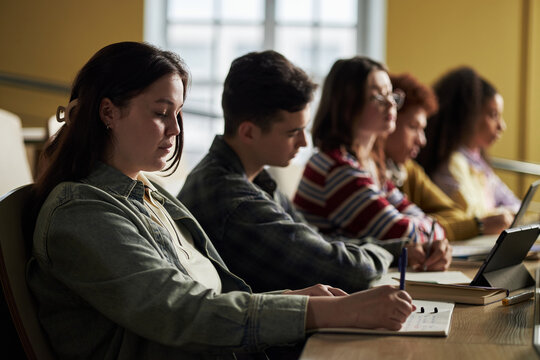 Polyethnic group of university students sitting in classroom taking notes and using laptop, young adults and teenagers focusing on studying during lecture, side view of diverse group - Powered by Adobe