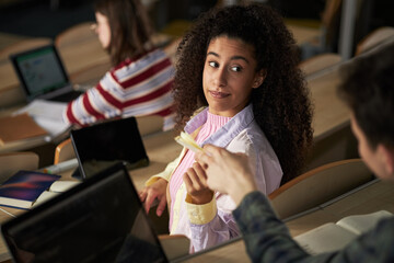 Young Black woman sitting in university lecture hall turning to look at Caucasian group mate offering pen, both surrounded by laptops and notebooks, studying together