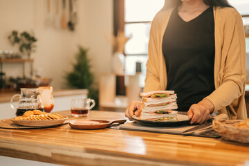 Close-up of a woman holding a plate with a stack of fresh sandwiches in a warm, modern kitchen....
