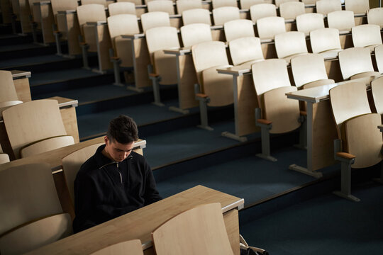 Young Caucasian man sitting alone in empty university lecture hall looking down at desk, appearing focused and thoughtful, surrounded by rows of unoccupied wooden seats